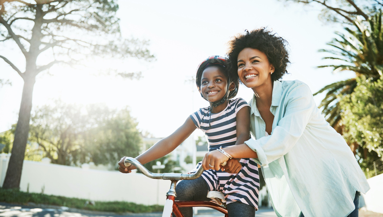 Mother joyfully helps her daughter learn to ride a bicycle on a sunny day, surrounded by trees and greenery.