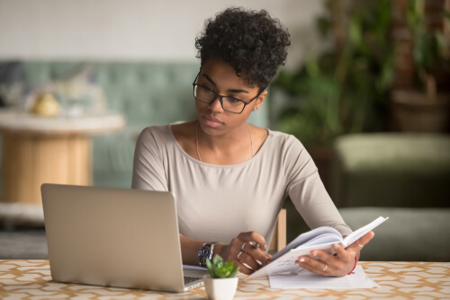 Person reading a book while using a laptop at a table with a small plant, focused on work.