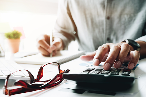 Person using a calculator and writing in a notebook, with glasses nearby, suggesting a focus on financial work.