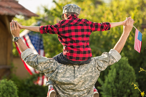 Child in red plaid shirt on adult's shoulders, holding small American flags, with larger flags in the background.
