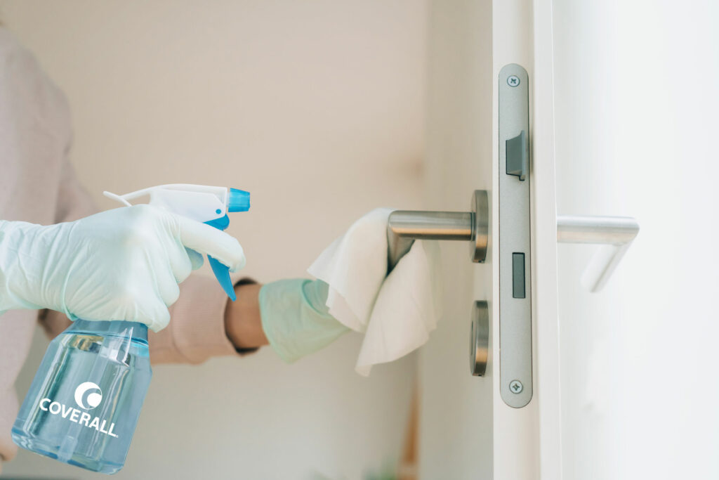 Gloved hands cleaning a door handle with a spray bottle and cloth for disinfecting.