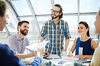 Team meeting in a bright office. Five people discuss and review documents at a table, smiling and engaging collaboratively.