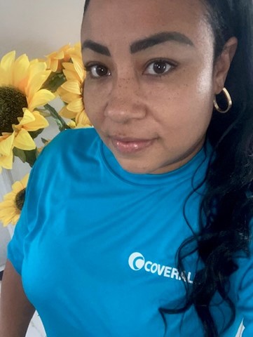 Person in a blue "Coverall" shirt with sunflowers in the background, looking into the camera.