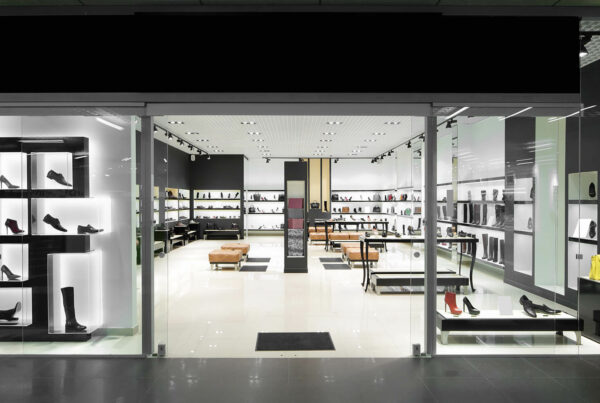 Modern shoe store with illuminated displays, featuring a variety of footwear on shelves and tables, viewed through glass door