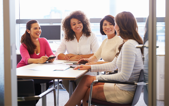 Four women in a meeting, seated around a table, smiling and engaged in conversation. Bright office setting with large windows