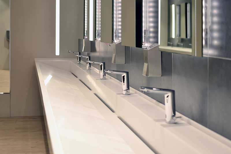 Modern public restroom with long white sink and multiple automatic faucets.