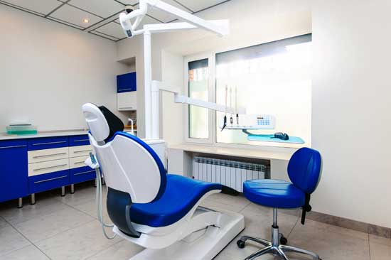 Dental office with a blue dental chair, equipment, and cabinets, lit by a large window.