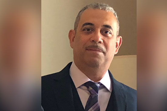 A man in a suit and tie, standing indoors with a neutral expression.