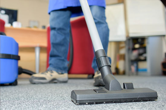 Person vacuuming office carpet with a vacuum cleaner, wearing blue jeans and work shoes. Office furniture is in the backgroun