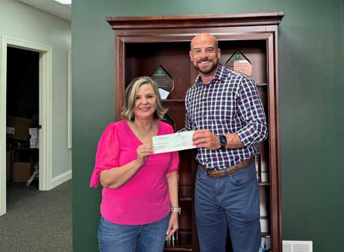 Two people smiling and holding a check in an office with a bookshelf in the background.