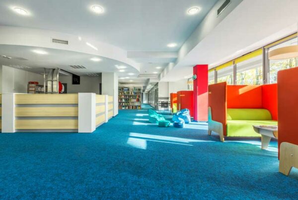 Modern library interior with blue carpet, colorful seating booths, bookshelves, and large windows allowing natural light.