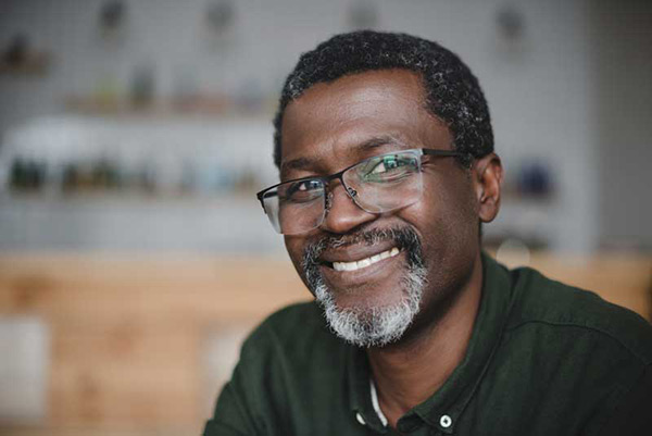 Smiling man with glasses and beard, sitting indoors, wearing a dark shirt.