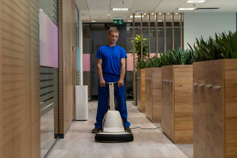 Person cleaning an office corridor with a floor polisher, surrounded by potted plants and wooden partitions.