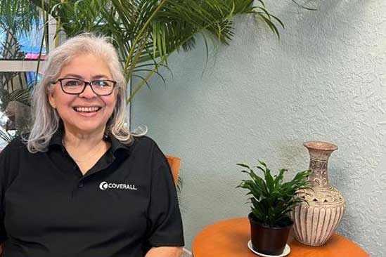 Smiling person in a black "COVERALL" shirt, seated beside a small plant and vase on an orange table, with greenery behind.