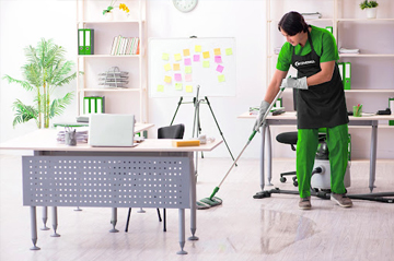 Person mopping an office floor near a desk and whiteboard in a well-organized space with shelves and plants.