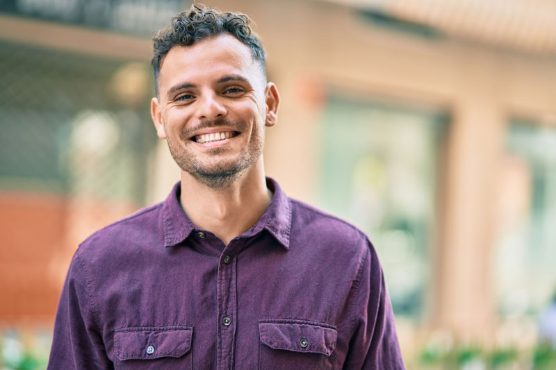 Smiling man in a purple shirt stands outdoors with a blurred city background.