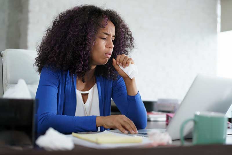 Woman in a blue sweater using a laptop at a desk, holding a tissue and appearing unwell.