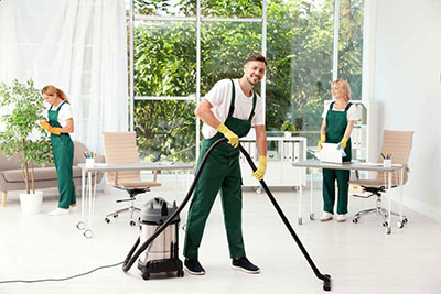 Cleaning crew in uniform, vacuuming and organizing a bright office space with large windows and greenery outside.