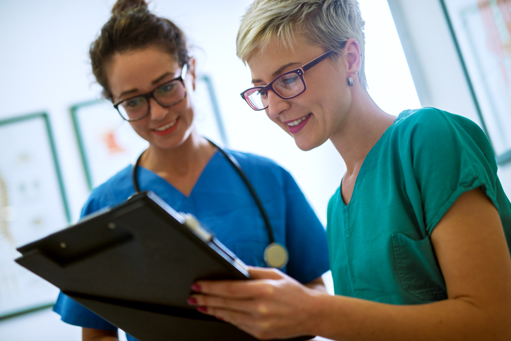 Two healthcare professionals in scrubs review a clipboard together in a medical office.