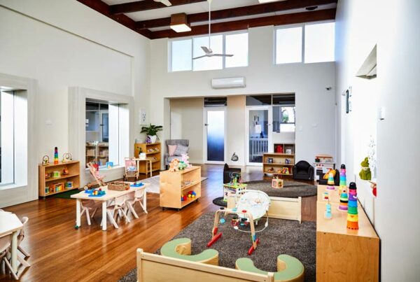 Bright daycare room with children's furniture, toys, and books on shelves and tables, featuring wood floors and large windows