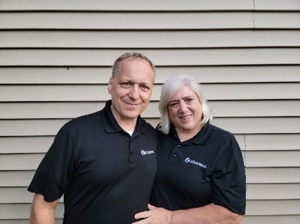 Two smiling people in matching black polo shirts stand together in front of a beige siding wall.