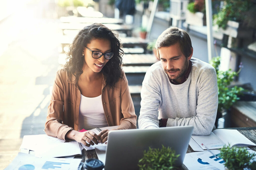 Two people reviewing documents and working on a laptop at an outdoor café table, with charts and plants visible.