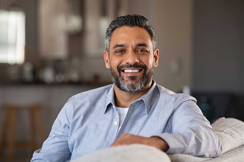 Smiling man in a light blue shirt sits on a couch in a cozy living room environment.