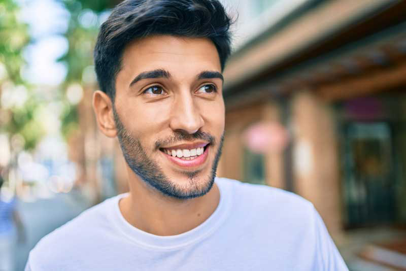 Smiling man outdoors with blurred city background, wearing a white shirt.
