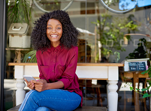 Smiling person in a red shirt sits outdoors on a bench, holding a smartphone, with plants and decor in the background.