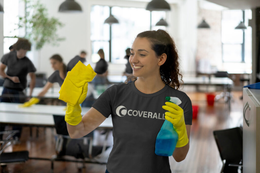 Person cleaning a glass surface with a yellow cloth and spray bottle, wearing a gray shirt and gloves in a bright room.
