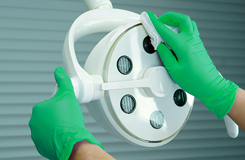 Person wearing green gloves cleans a round surgical lamp with a cloth.