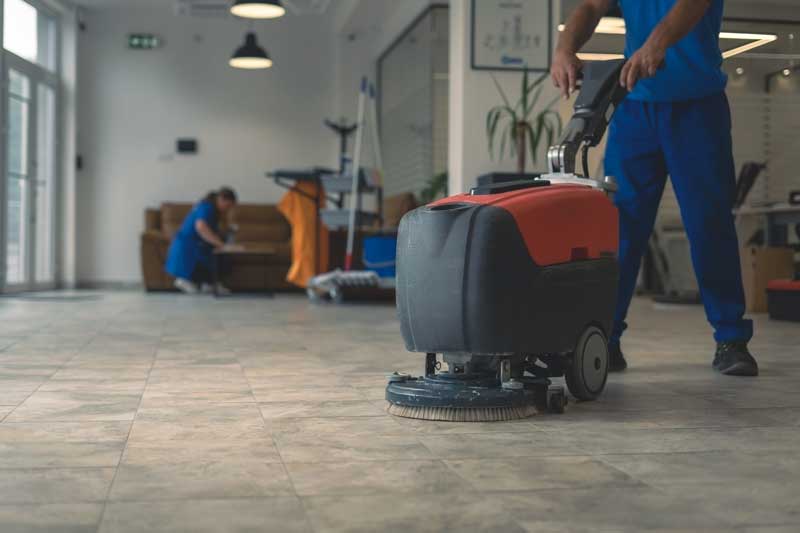 Worker in blue uniform using a floor scrubber in a large, tiled room with cleaning equipment in the background.