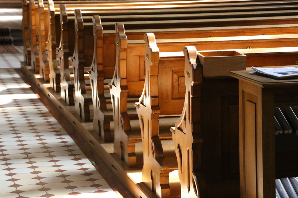 Wooden church pews with decorative end panels, sunlit, beside a tiled floor and a small wooden table holding papers.