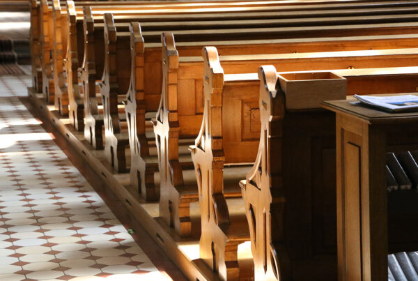 Wooden church pews with decorative end panels, sunlit, beside a tiled floor and a small wooden table holding papers.