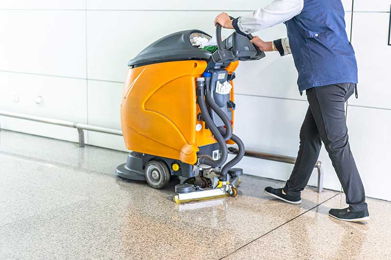 Person operating an orange floor-cleaning machine in a hallway.