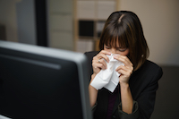 Person in office blowing nose with tissue, sitting in front of a computer monitor.