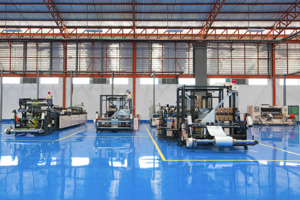 Industrial warehouse interior with packaging machinery on a shiny blue floor, under a high metal roof.
