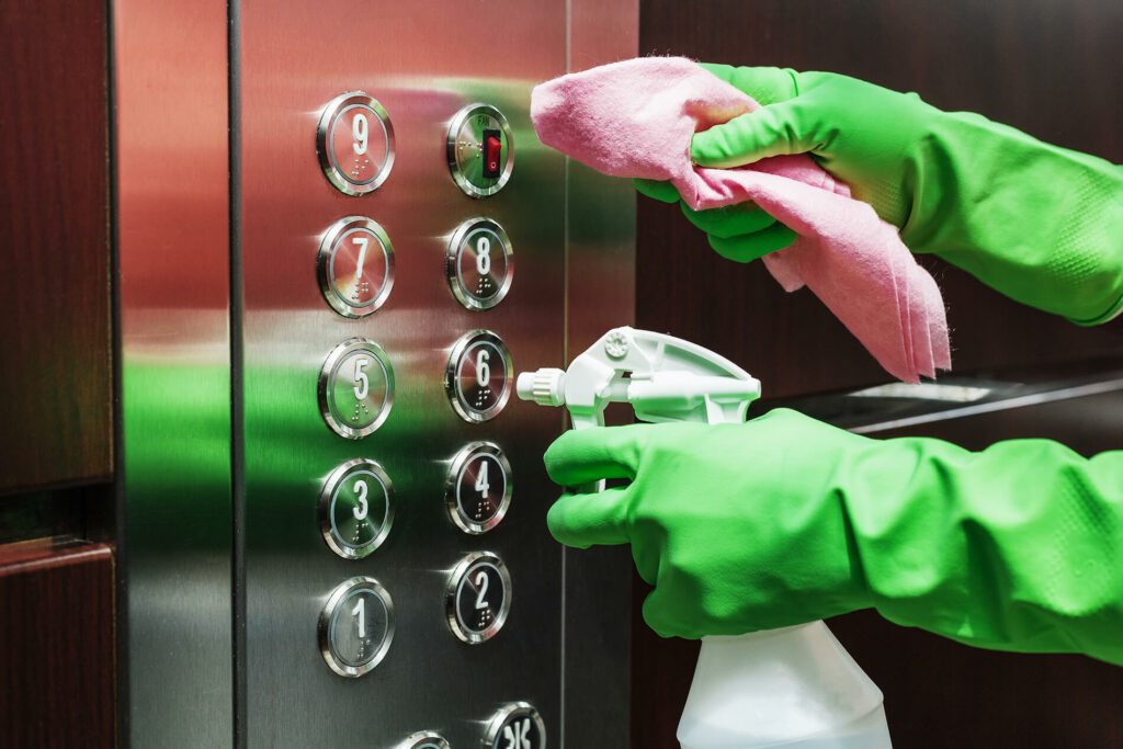 Person in green gloves cleaning elevator buttons with spray and cloth.