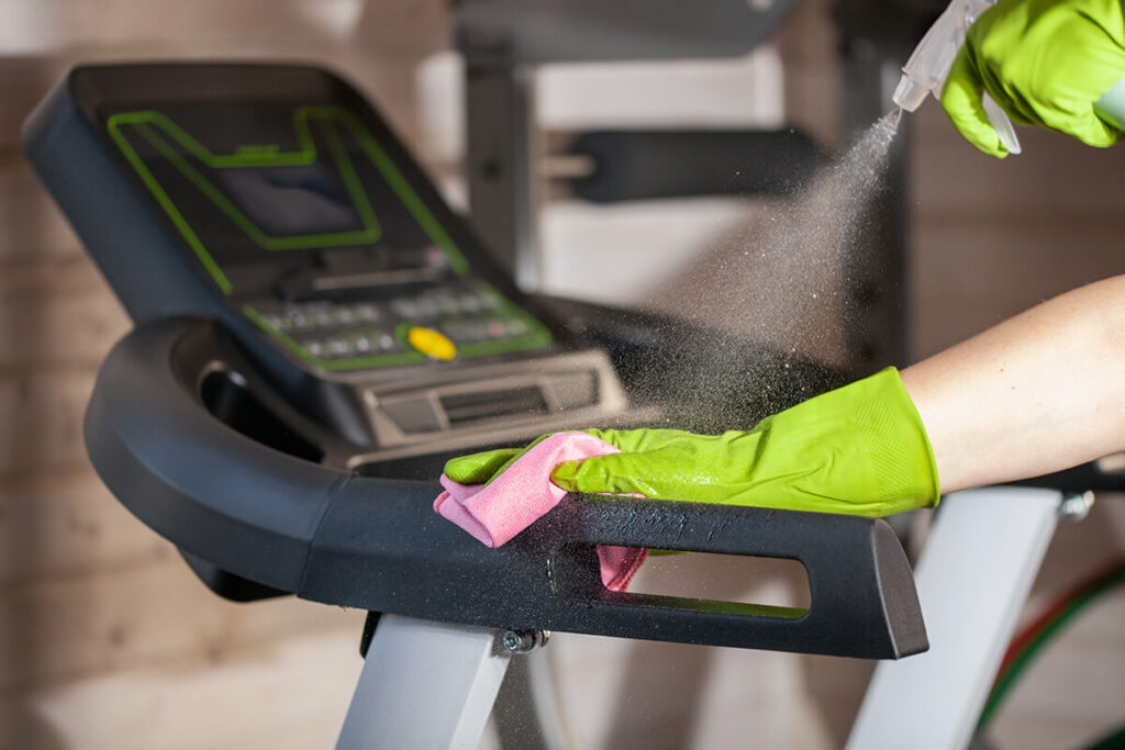 Person wearing gloves cleans treadmill with spray and cloth.