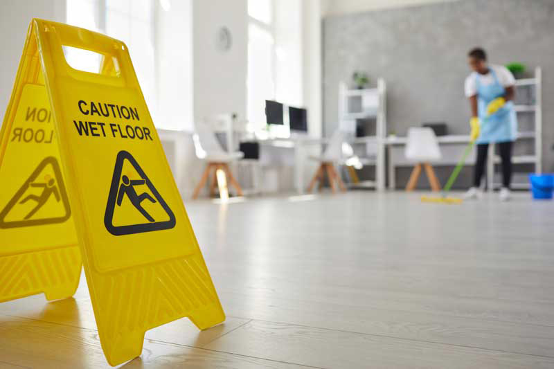 Yellow "Caution Wet Floor" sign with a person mopping the floor in a bright office.