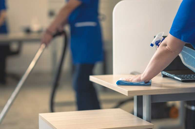 Person cleaning desk with cloth and spray bottle, another vacuuming in background. Office setting.