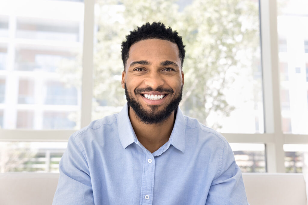 Smiling man in a blue shirt sitting indoors with large windows in the background.
