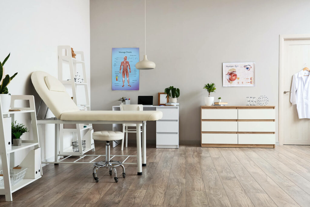 Medical office with examination table, stool, posters, and cabinets. White walls and wooden flooring add a modern touch.