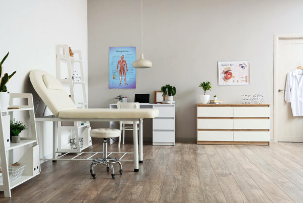 Medical office with examination table, stool, posters, and cabinets. White walls and wooden flooring add a modern touch.