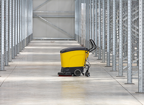 Yellow floor scrubber in an empty warehouse aisle with metal shelves on both sides.