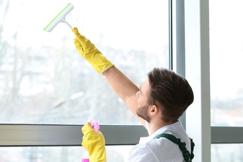 Person in yellow gloves cleaning a window with a squeegee, holding spray bottle, daylight outside.