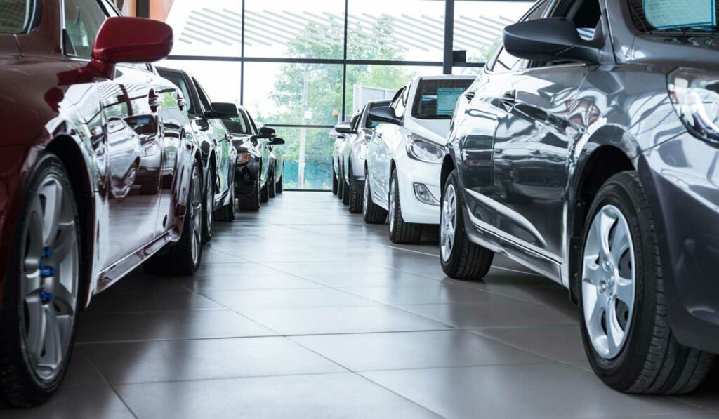 Cars are lined up inside a dealership showroom with a large window in the background.