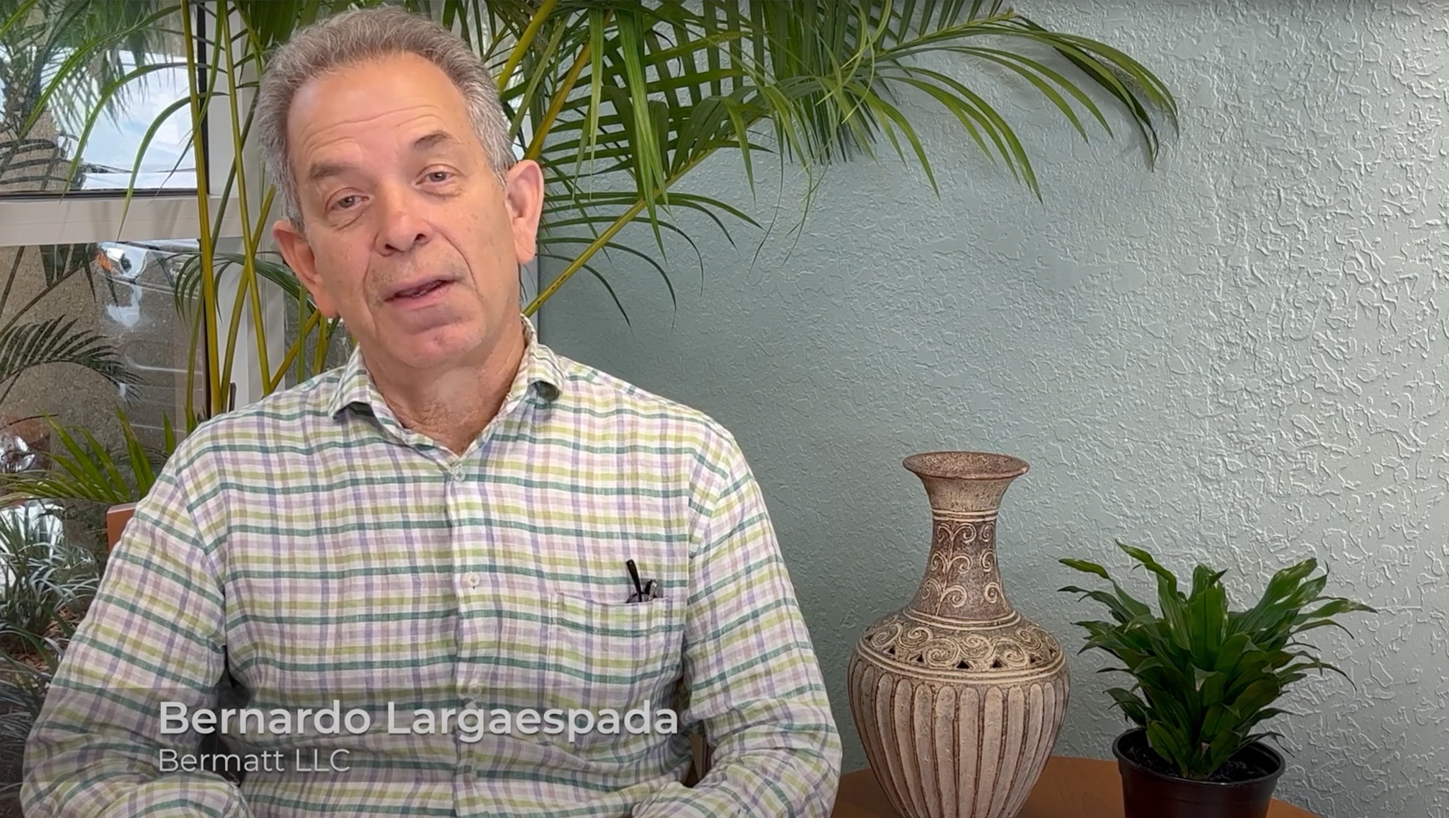 Man in plaid shirt seated by plants and a decorative vase, against a textured wall background.