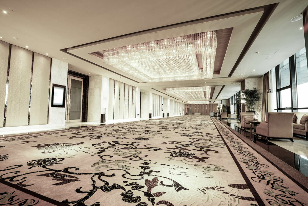 Luxurious hotel lobby with patterned carpet, chandeliers, and seating areas. Neutral tones and natural light fill the space.