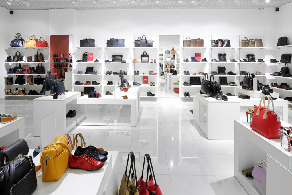 Modern shoe store interior with white shelves displaying various shoes and handbags under bright lighting.